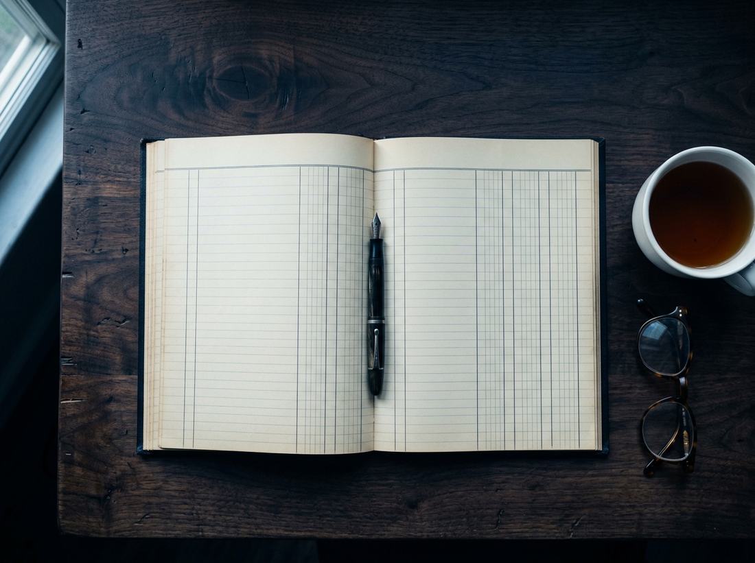 An accounting ledger open at ruled columns on a dark desk, with a pen and reading glasses